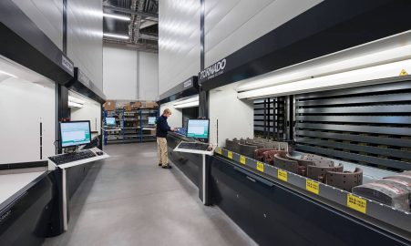 An industrial warehouse featuring automated "Tornado" vertical lift modules, with a worker operating a control terminal and trays of mechanical parts visible.