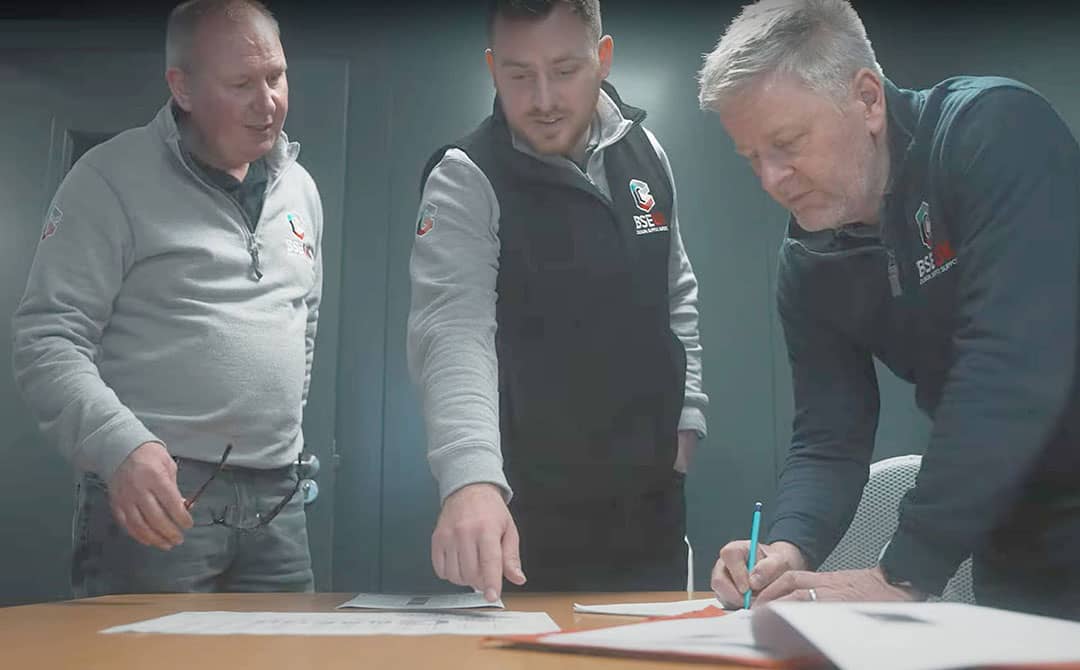Three BSE UK team members, representing a SEMA-approved team, collaborating over project plans and documentation at a desk.