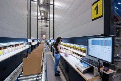 A warehouse employee working at a computerized order-picking and packing station along a conveyor line.