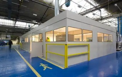 A modular white warehouse office with large glass windows, protected by yellow safety barriers.