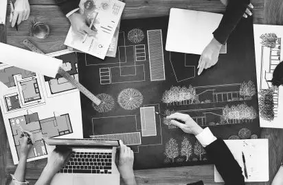 A black and white overhead view of hands collaborating on architectural blueprints and project plans on a large desk.