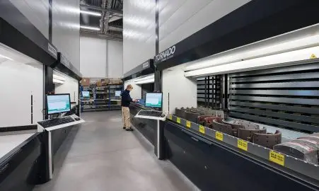 An industrial warehouse featuring automated "Tornado" vertical lift modules, with a worker operating a control terminal and trays of mechanical parts visible.