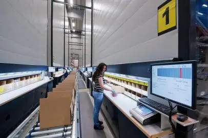 A warehouse employee working at a computerized order-picking and packing station along a conveyor line.